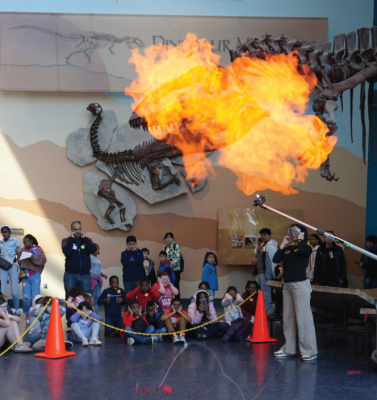 group of people sit on museum floor while fiery explosion happens overhead